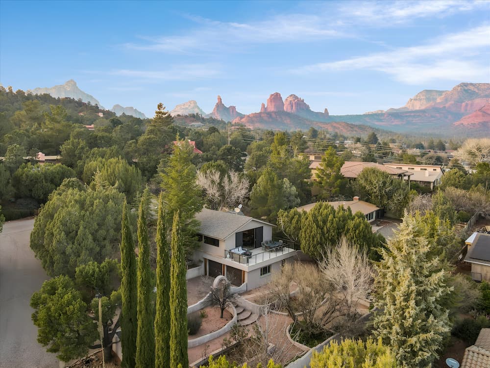 Aerial view of desert home surrounded by trees with red rock formations in distance