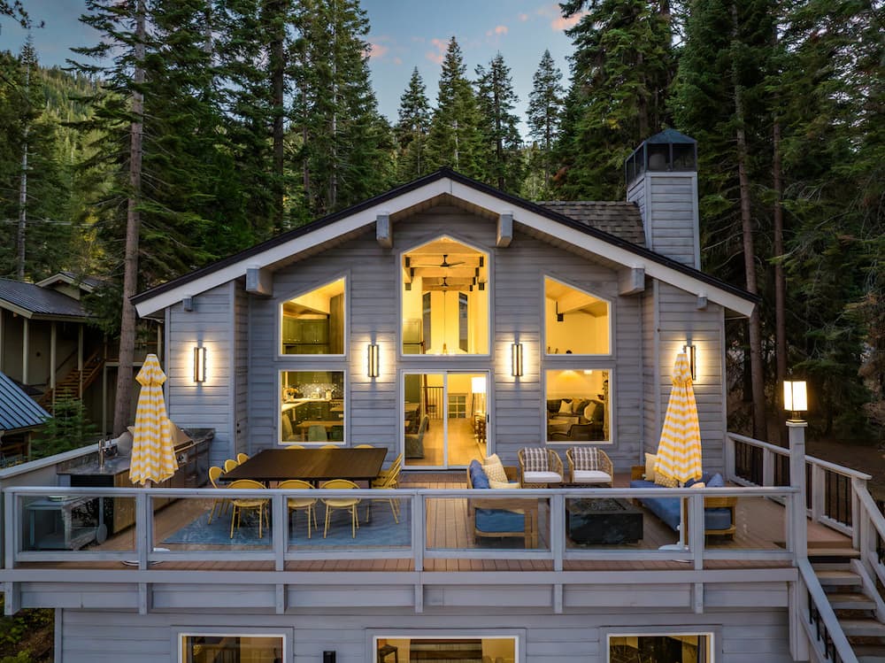 Mountain cabin with large windows and wraparound deck dining area at dusk