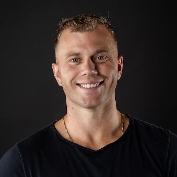 Headshot of smiling man in black t-shirt with gold chain against dark background