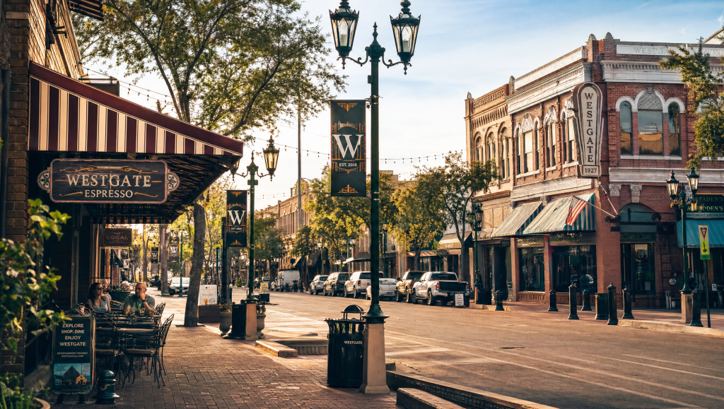 A sunny street scene in Historic Downtown Glendale, Arizona, featuring storefronts like Westgate Espresso with striped awnings, outdoor seating, mature trees, and historic brick buildings along a quiet road.